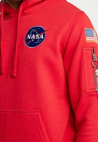 Punainen huppari, jossa sininen NASA-logo ja amerikkalaisen lipun merkki. Hupussa on etutasku ja vetoketjullinen hihatasku.