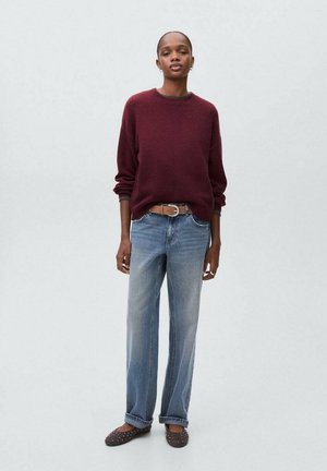 Bordeaux trui, blauwe losse jeans, bruine riem en bruine schoenen met studs. Model staat tegen een lichtgrijze achtergrond.