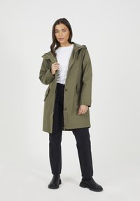 Oliwkowa wodoodporna kurtka z kapturem, z zamkiem błyskawicznym z przodu oraz zapięciem na napy; w komplecie z czarnymi dżinsami i botkami za kostkę.