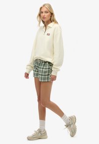 Sudadera de color amarillo claro con cuello de cremallera y parche con logo, combinada con pantalones cortos de cuadros verdes. El modelo lleva zapatillas deportivas claras y calcetines blancos.