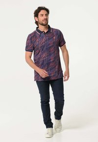 Poloshirt met een donkerblauwe basis en een levendig bladmuster in tinten van rood en oranje, aangevuld met contrasterende accenten op de kraag en mouwen.