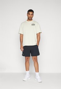 Světle béžové oversized tričko s černým grafickým potiskem, spárované s tmavě námořnickými šortkami. Model nosí bílé tenisky a ponožky do délky kotníků.