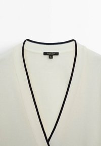 V-neck sweater in cream with a textured knit pattern and contrasting black trim at the neckline. Label visible inside collar.