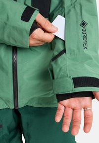 Een witte kaart wordt met de hand in de ritszak op de borst van een groene Gore-Tex jas gestoken, die over een gelaagde zwarte manchette wordt gedragen.