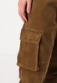 Bruine corduroy cargobroek met een zijvak, ribbeltextuur en een getailleerd ontwerp. De stof heeft een zachte afwerking en zichtbare stiksels.