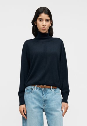 Πλεκτό μπλε σκούρο πουλόβερ με ζιβάγκο από απαλό υλικό, με υφασμένη μπροστινή επιφάνεια. Συνδυασμένο με ανοιχτό μπλε, ψηλόμεσα τζιν παντελόνια και καφέ ζώνη.