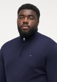 Granatowy sweter z zamkiem w kolorze navy, o gładkiej teksturze dzianiny. Na piersi znajduje się małe logo w kolorach czerwonym, białym i niebieskim. Prosty, nowoczesny design.