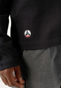 Camicia nera a maniche lunghe in cotone con patch rotondo del logo JOTT che presenta una figura seduta e un accento rosso in fondo, abbinata a pantaloni in tessuto grigi.