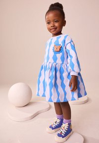 Plava i bijela haljina s uzorkom valova i dugim rukavima, s aplikacijom u obliku lava na prsima, upotpunjena šarenim tenisicama.