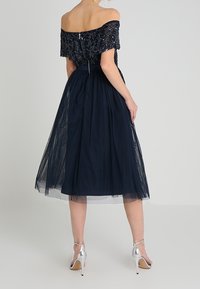 Námořnická off-shoulder šaty s vyšívaným živůtkem, upraveným pasem a středně dlouhou tylovou sukní. Stříbrné vysoké podpatky dokončují vzhled.