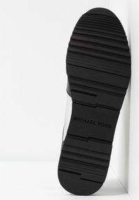 Semelle en caoutchouc noire d'une chaussure avec des crampons horizontaux surélevés et "Michael Kors" embossé dans la partie centrale.