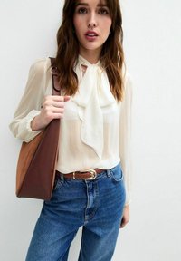 Light beige blouse with a tie neck, textured fabric; paired with blue denim jeans and a brown leather shoulder bag. Bronze belt buckle detail.