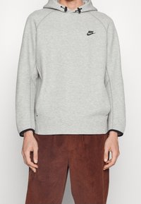 Persona vistiendo una sudadera con capucha de Nike en gris claro con el logo negro en el pecho y pantalones de pana marrones, de pie contra un fondo liso.