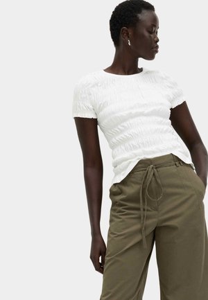 Vrouw met kort haar, gekleed in een witte textuur korte mouw top en olijfgroene broek met trekkoord, staand met één hand in de zak.