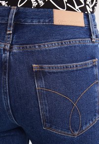 Tummansiniset farkut, joissa on sileä pinta, varustettuna nahkaleimalla, jossa lukee "Calvin Klein Jeans", ja erottuvilla ompeluominaisuuksilla taskuissa.