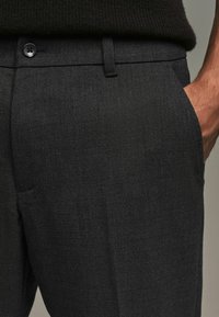 Black textured trousers featuring a smooth fabric, front pockets, and a button closure, shown with a hand resting on the pocket.