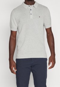 Ljusgrå bomullspolo med ribbad textur, tre knappar och en liten logotyp på bröstet. Kort ärm och klassisk kragdådesign.