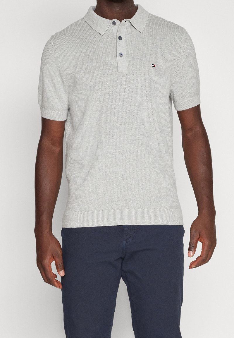 Ljusgrå bomullspolo med ribbad textur, tre knappar och en liten logotyp på bröstet. Kort ärm och klassisk kragdådesign.