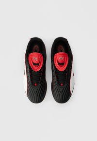 O pereche de teniși Puma negri, albi și roșii văzuți dintr-o perspectivă de sus pe un fundal alb, având limbi roșii proeminente cu logo-uri Puma.