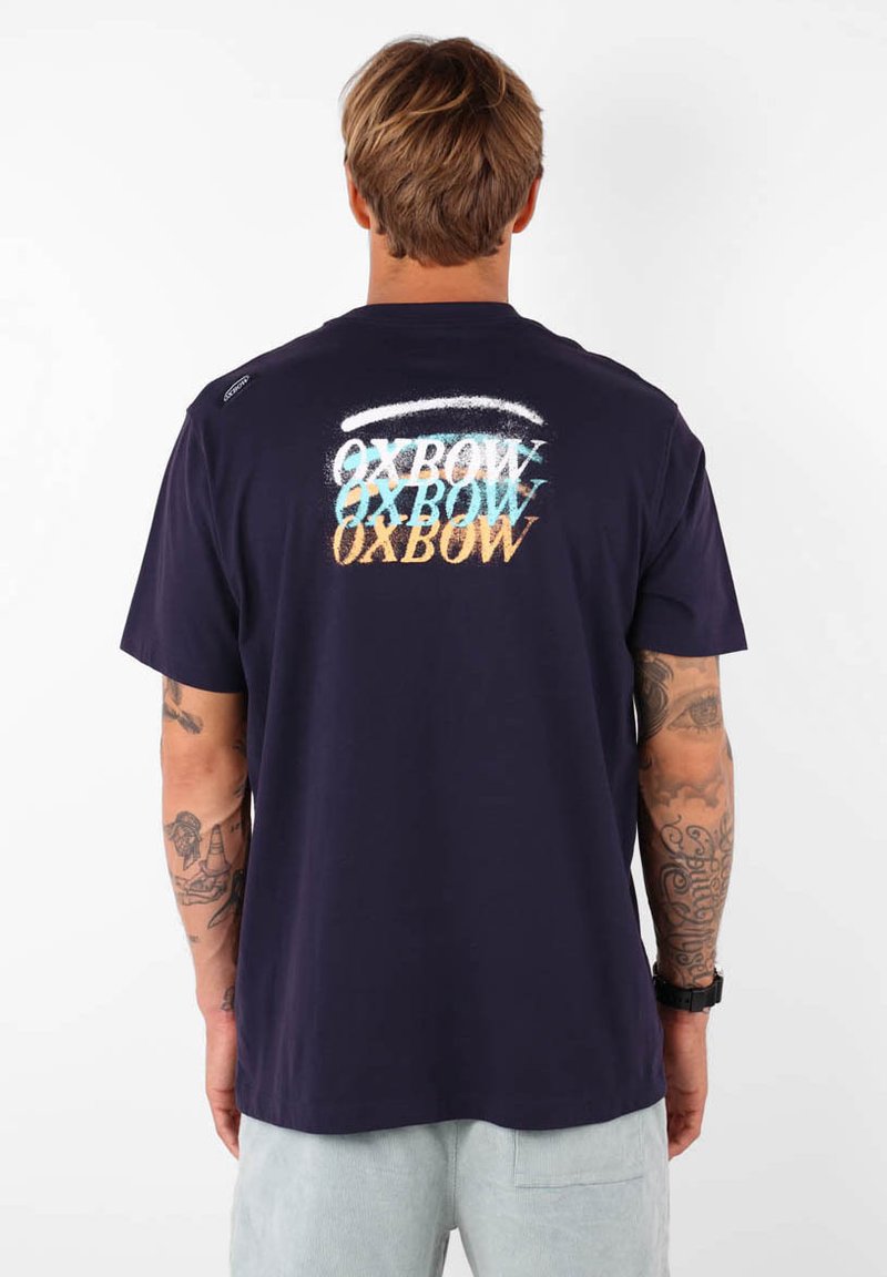 Camiseta de algodón azul marino con diseño de texto "OXBOW" multicolor en la parte posterior, manga corta y escote redondo.