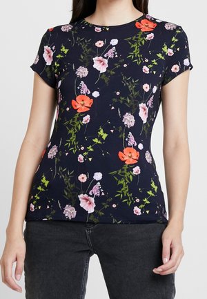 Kobieta w dopasowanym granatowym topie z krótkim rękawem z kolorowym kwiatowym wzorem i czarnych dżinsach, stojąca z rozluźnionymi ramionami przy bokach.