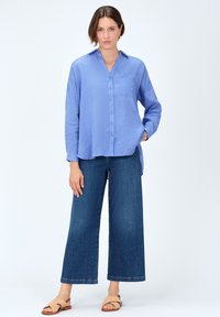 Camicia azzurro chiaro a maniche lunghe con colletto e tasca, abbinata a pantaloni in denim scuri a gamba larga e sandali intrecciati beige.