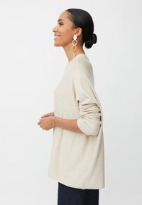 Beiger Strickpullover mit lockerer Passform, weiten Ärmeln und gerippten Bündchen. Kombiniert mit großen goldenen Creolen, seitliche Ansicht.