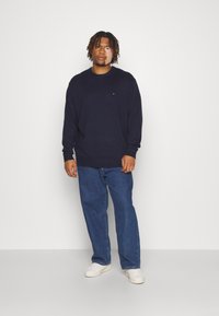 Navy knitted sweatshirt with long sleeves, round neckline, and small logo detail above the left chest, paired with light blue denim jeans and white sneakers.
