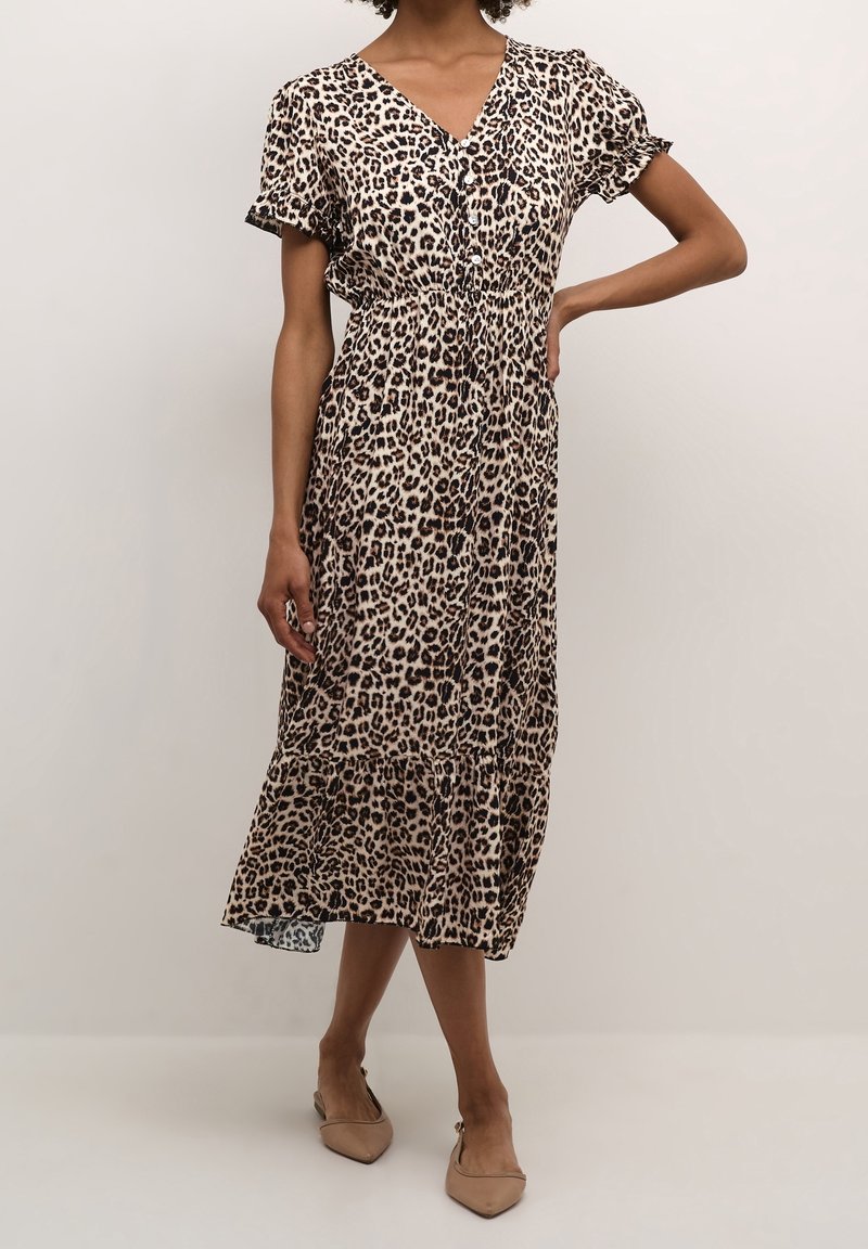 Frau in einem knielangen Kleid mit Leopardenmuster und kurzen Rüschenärmeln, kombiniert mit beigen spitzen Slingback-Pumps, steht vor einem schlichten Hintergrund.