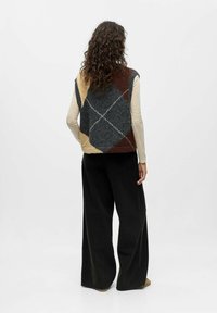 Strickweste mit einem Argyle-Muster in Grau, Braun und Beige. Getragen über einem hellen Langarmshirt, kombiniert mit weiten schwarzen Hosen.