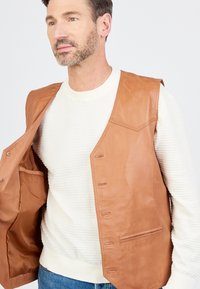 Bruine leren gilet met een gladde textuur, met een knoopsluiting aan de voorkant en twee zijzakken, gedragen over een witte geribbelde trui.