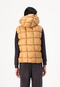 Gilet imbottito giallo senape con motivo a quadri e cappuccio oversize, indossato sopra un outfit scuro a coste, senza hardware visibile.