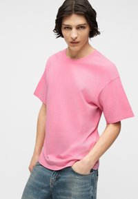 Νεαρό άτομο με μεσαίου μήκους σκούρα μαλλιά, φορώντας ένα άνετο ροζ T-shirt και μπλε τζιν, που ποζάρει με το ένα χέρι στην τσέπη μπροστά σε λευκό φόντο.