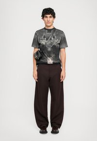 Black graphic t-shirt with a distressed design, loose brown pants, and a small black bag with a shoulder strap. Dark shoes complete the look.
