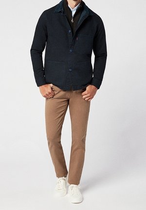 Mann trägt eine dunkelblaue Jacke mit Knöpfen über brauner Hose und weißen Turnschuhen, steht vor einem schlichten hellen Hintergrund.