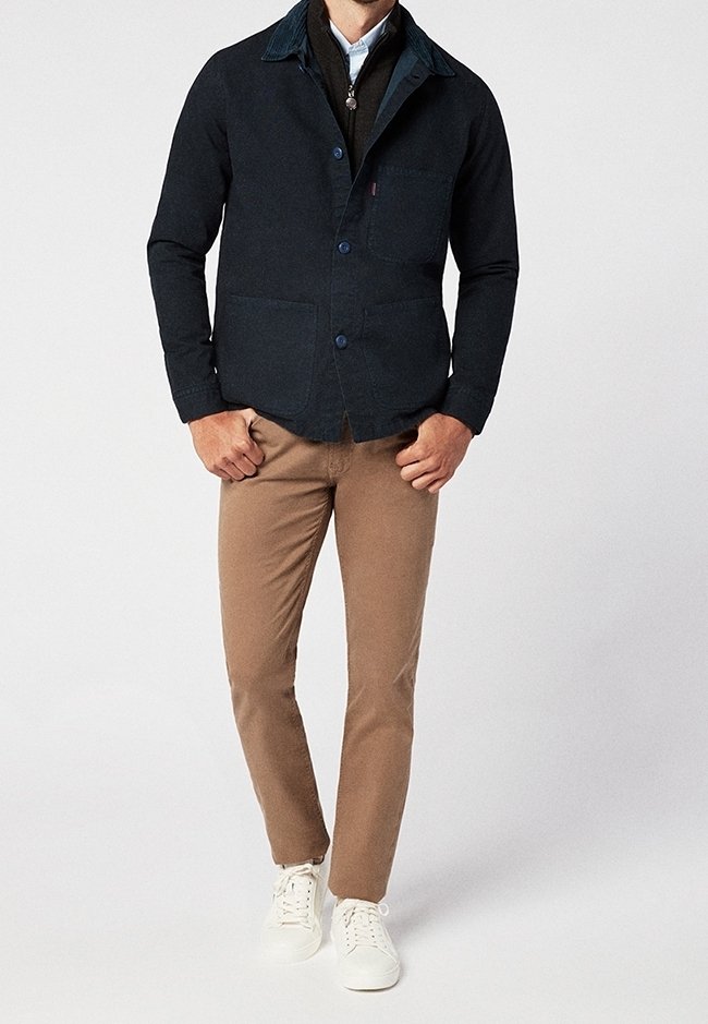 Homme portant une veste boutonnée bleu marine foncé sur un pantalon marron et des baskets blanches, debout devant un fond clair uni.
