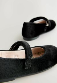 Scarpe Mary Jane in suede nero con punta arrotondata, cinturino regolabile e soffice fodera interna. Suole piatte e dettagli sottili sul cinturino.
