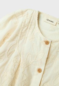 Cremfarbene Bluse mit floralem Stickdesign und detaillierten Blatt- und Blumenmustern. Mit Holzknöpfen an der Vorderseite vollendet.