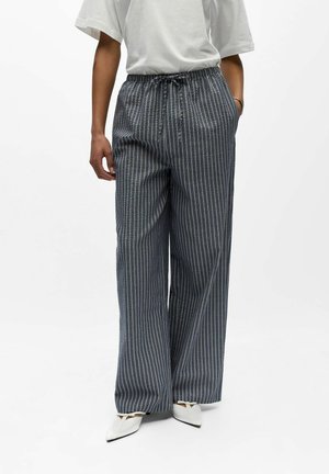 Osoba ubrana w luźne, wysokie w talii, pionowo paskowane szare spodnie ze sznurkiem, białe spiczaste buty oraz prostą białą koszulkę z krótkim rękawem.