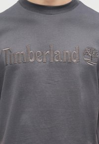 Σκούρα γκρι φούτερ με ανάγλυφο κεντημένο κείμενο "Timberland" και λογότυπο δέντρου στο στήθος, που φοριέται από ένα άτομο.