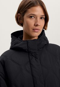 Veste matelassée noire avec un col haut et un motif de couture complexe. Fabriquée à partir d'un matériau lisse et léger avec une finition mate.