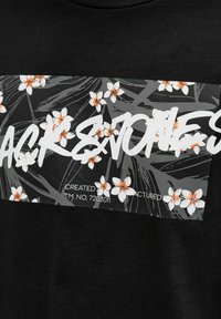 Sort t-shirt med et grafisk hvidt blok med blomstermotiv og tekst. Blomsterne er hvide med orange midte, sat mod en mørk baggrund.