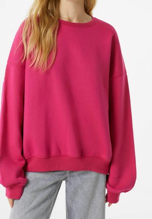 Femme portant un sweat-shirt rose vif oversize à col rond avec manches longues et un jean gris clair, visible des épaules aux hanches.
