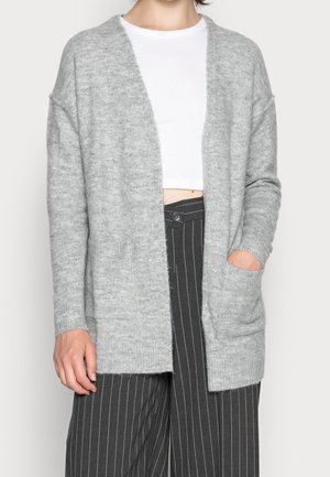 Personne portant un cardigan gris clair ouvert sur un crop top blanc et un pantalon noir taille haute à rayures larges et jambes larges.