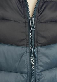 Giubbotto imbottito con un design bicolore in grigio scuro e verde petrolio. Chiusura centrale con zip abbinata e dettagli di cucitura.