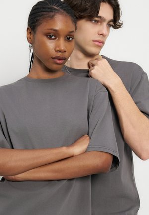 Ung svart kvinne med flettehår og ung hvit mann med bølgete hår står tett sammen med seriøse uttrykk, begge iført grå t-skjorter.