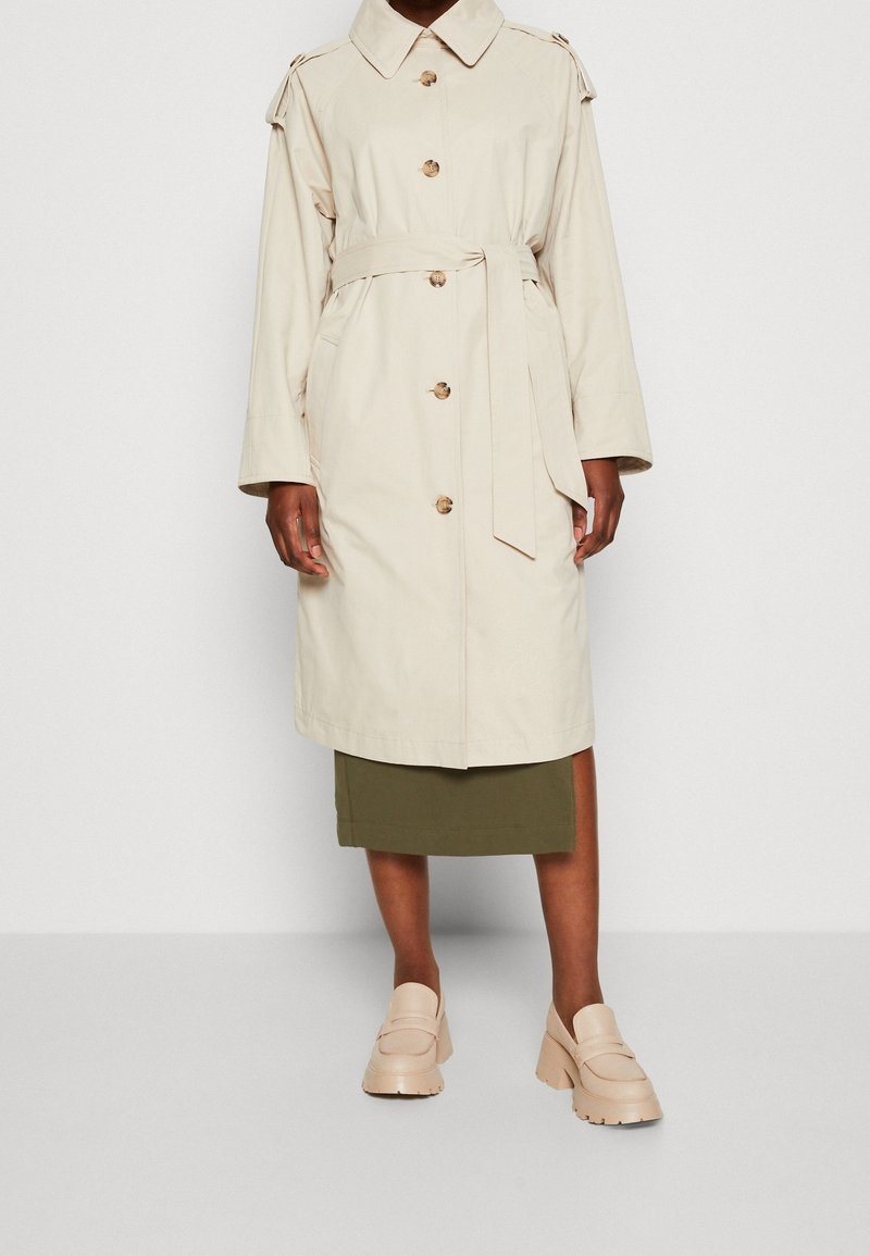 Persona vestida con un abrigo trench beige ceñido con cinturón sobre una falda verde oliva y mocasines de tacón grueso beige, de pie frente a un fondo liso.