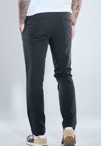 Man with tattooed arms wearing black slim-fit pants and white sneakers with beige accents, standing with hands in pockets.