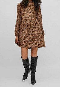 Kobieta ubrana w długą sukienkę w kolorze brązowym i czarnym w paski tygrysa, połączoną z czarnymi, wysokimi na kolana, obcasami, na jednolitym tle.