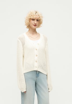 Osoba s kučeravými blond vlasmi má na sebe biely sieťovaný pletený sveter na gombíky cez biely top a svetlomodré rifle s vysokým pásom.